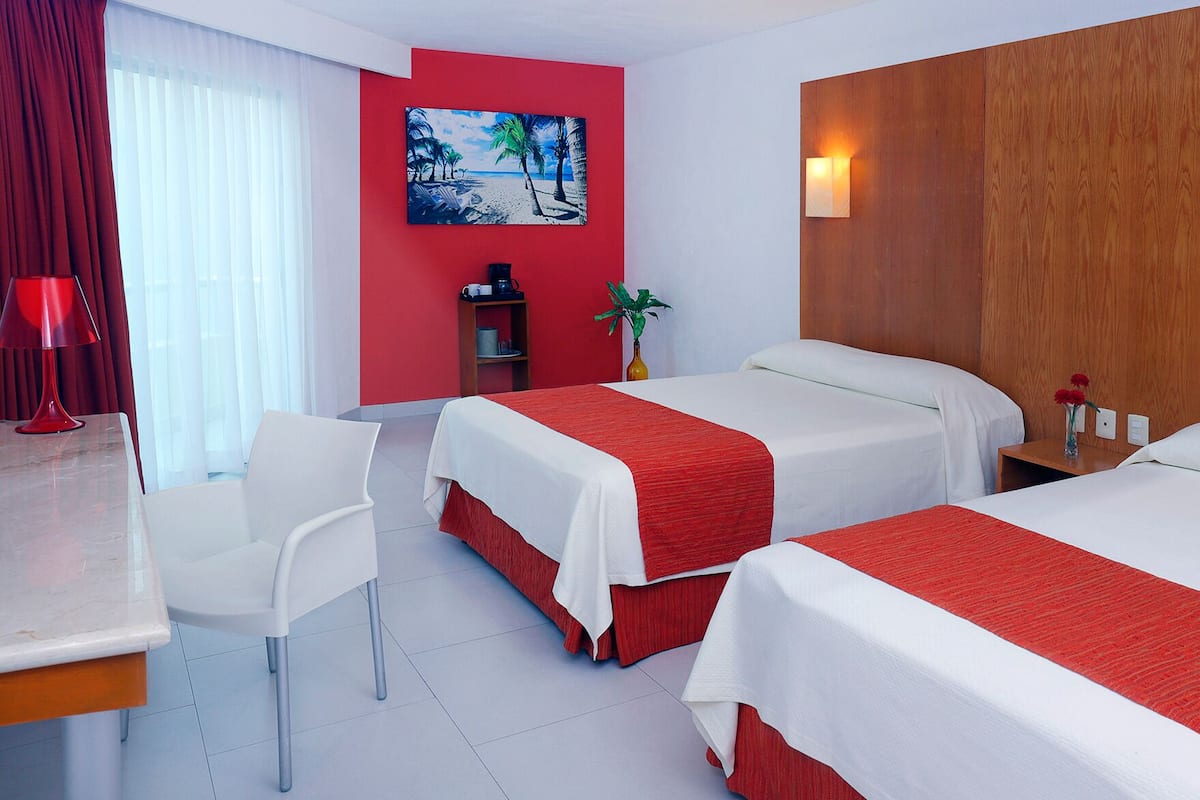 Habitación de hotel con dos camas, un escritorio, una silla, pared roja, un cuadro y paneles de madera.