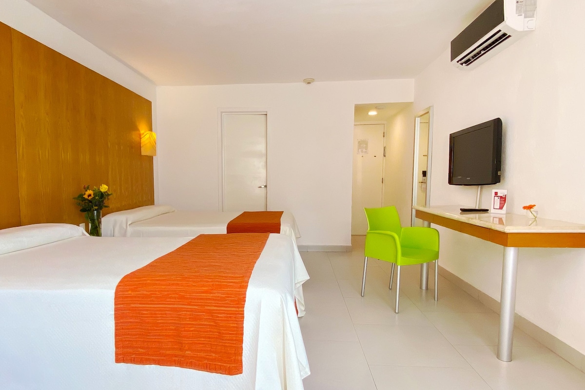 Une chambre d’hôtel avec deux lits, un bureau, une chaise, une télévision et une porte donnant sur une autre pièce.