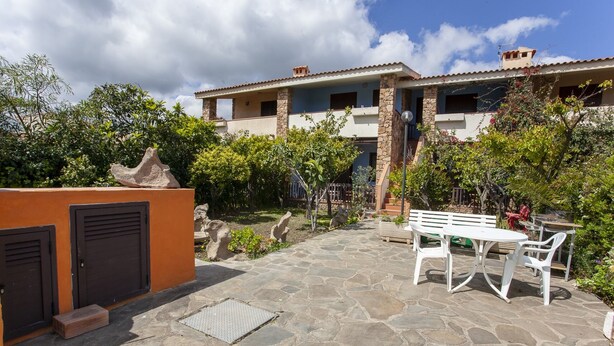A courtyard with a stone floor, a white bench, and a table. There are two buildings with balconies and a stone wall with two doors. - Residence La Cinta 7 1P (San Teodoro)