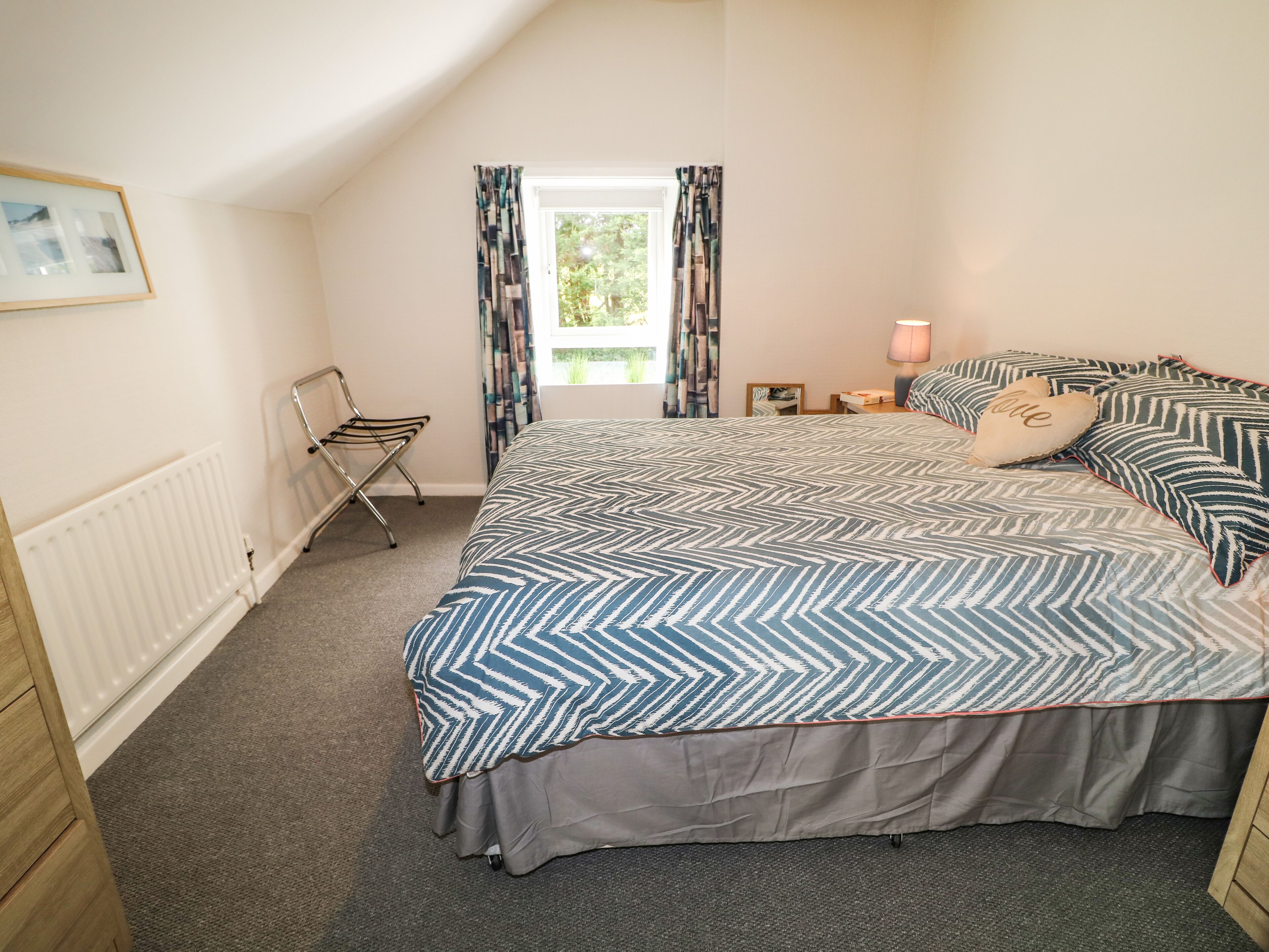A bedroom with a bed, a radiator, a chair, a window with curtains, and a nightstand with a lamp.
