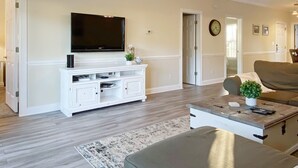 Una sala de estar con un televisor de pantalla plana montado en un mueble blanco, un sofá marrón, una mesa de centro con una planta y un tapete estampado.