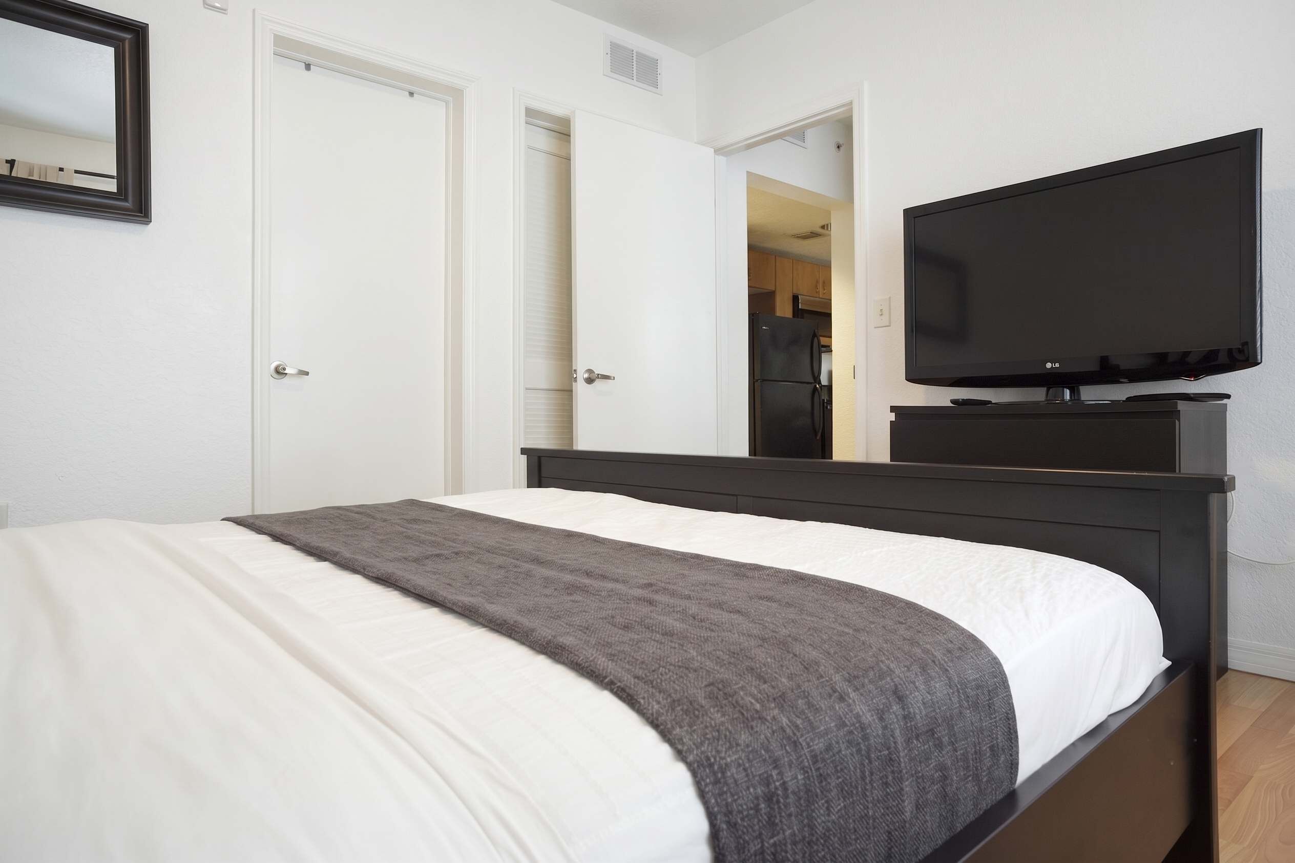 A bedroom with a bed, a television on a stand, and a door leading to a kitchen.