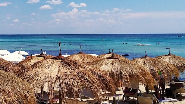 Ein Strand mit Reetsonnenstühlen und Liegestühlen, ein strahlend blauer Himmel und das Meer im Hintergrund.