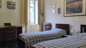 Ein Hotelzimmer mit zwei Betten, einem Schreibtisch, einem Sessel, einem Heizkörper und einem fenster mit Vorhängen.