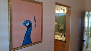 A hallway with a framed painting of a peacock, a bathroom with a sink and towel rack, and a door leading to another room. - Dark Horse Ranch · Single-Story Home on 5 Acres · Sleeps 8 · Dog-Friendly (Yucca Valley)
