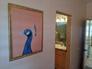 Ein Flur mit einem gerahmten Pfauengemälde, ein Badezimmer mit Waschbecken und Handtuchhalter, sowie eine Tür zu einem weiteren Zimmer.