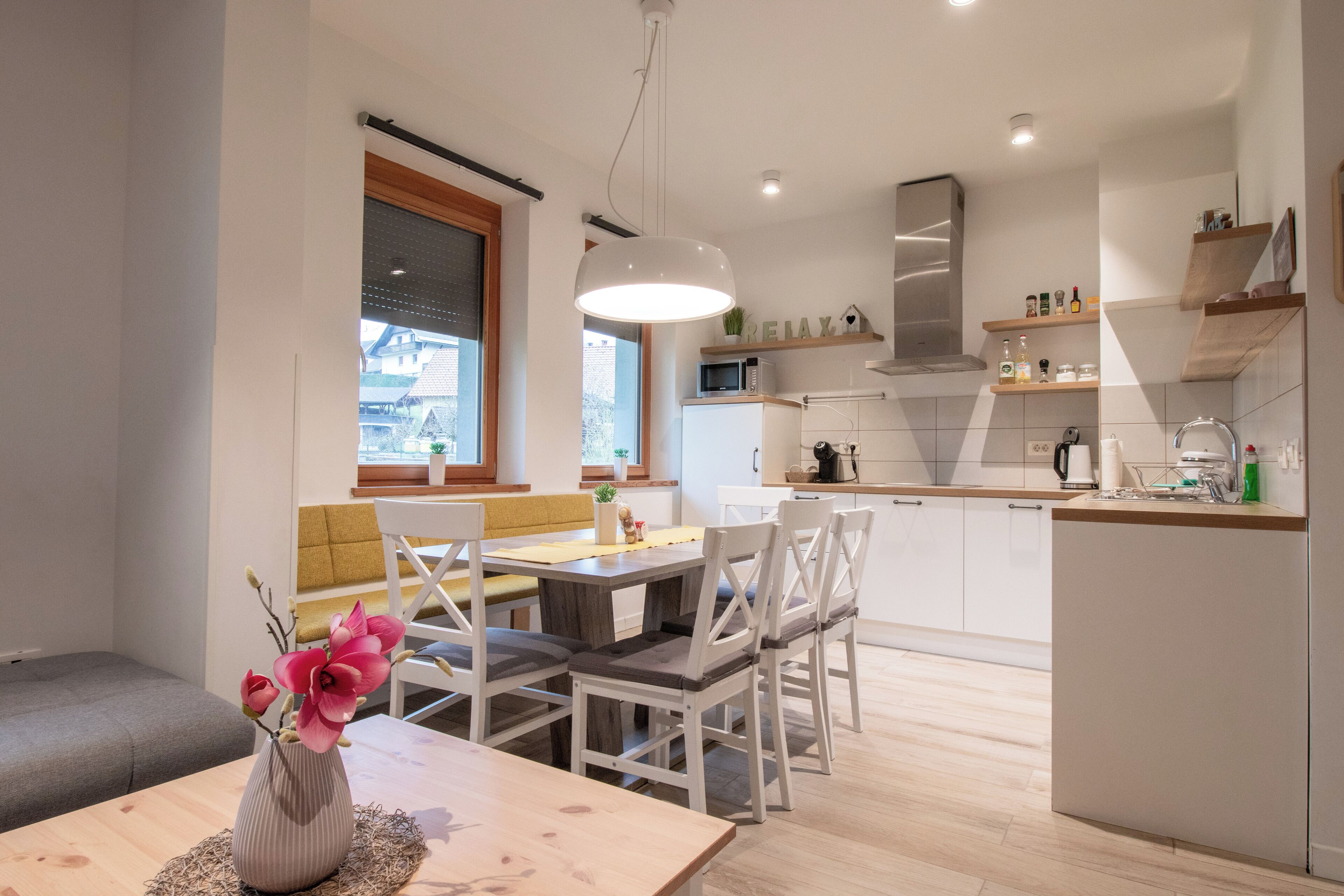 A modern kitchen with white cabinets, a dining area with a wooden table and chairs, and a view of the outside through a window.