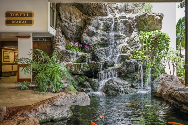 Cascada de rocas y estanque con peces koi, rodeado de plantas tropicales.