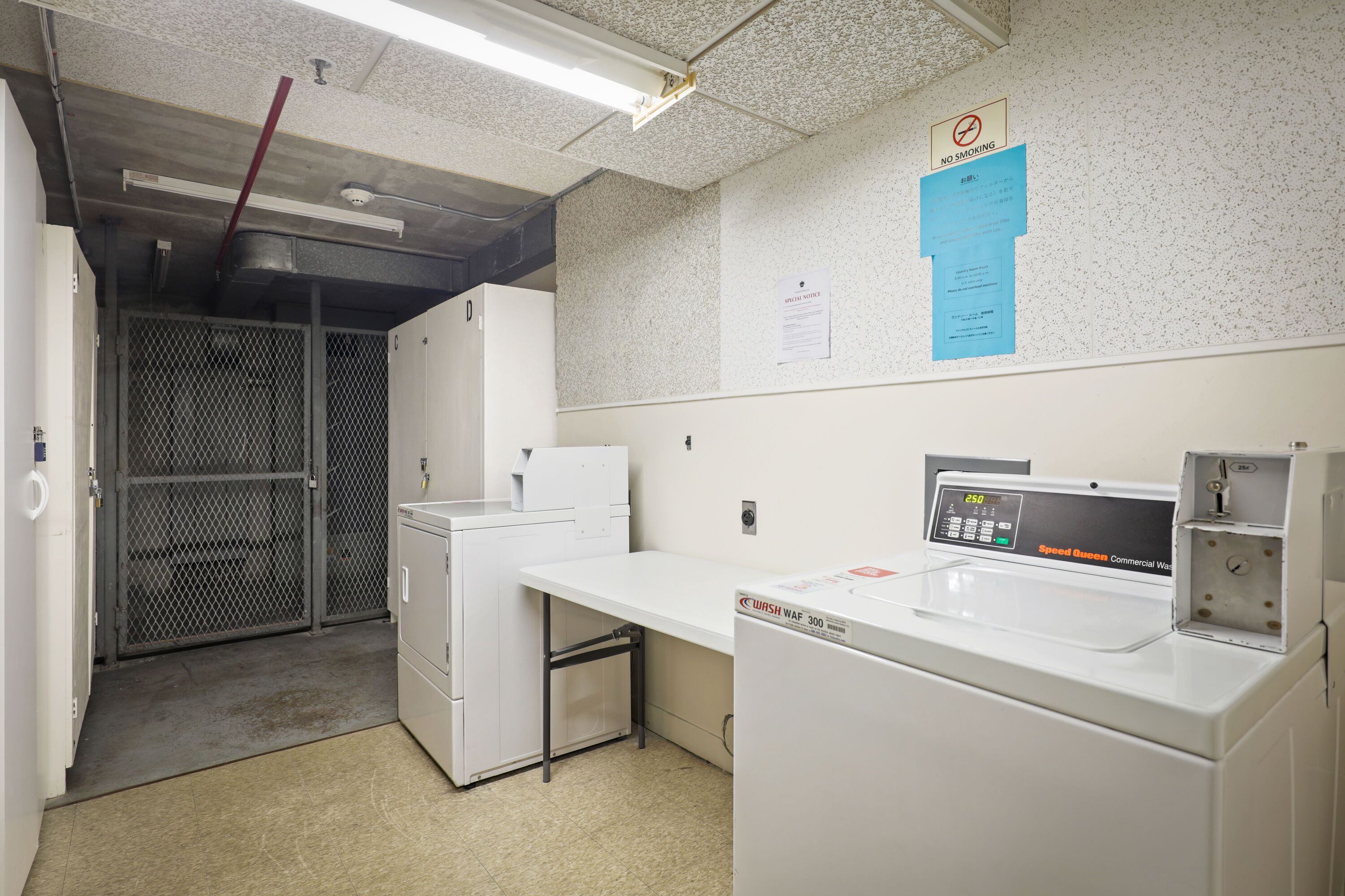 A laundry room with a commercial washer, a drying machine, and a control panel. There is a 'No Smoking' sign on the wall and a notice about laundry room hours.