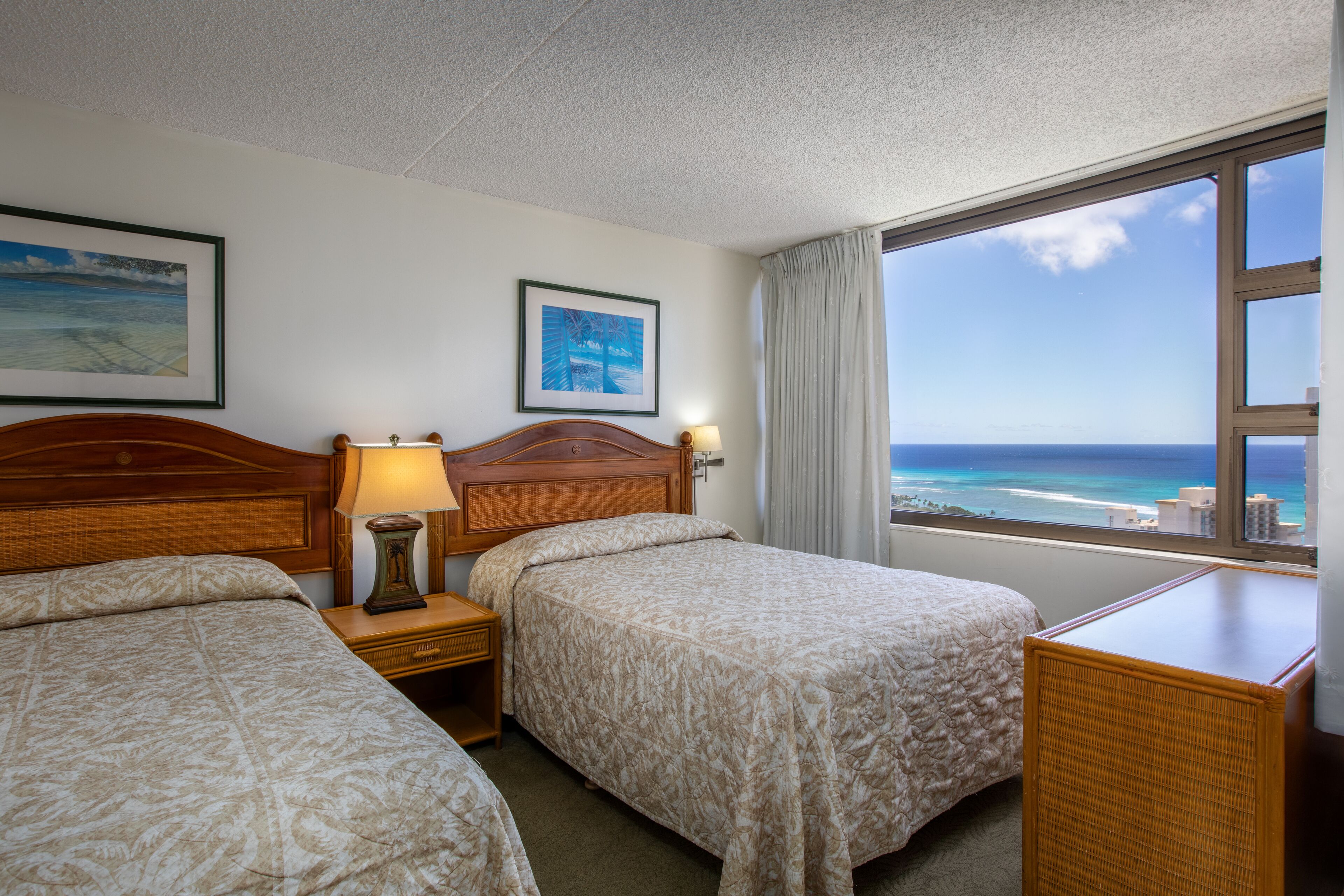 A hotel room with two beds, a wooden headboard, a nightstand with a lamp, a window with a view of the ocean, and framed pictures on the wall.