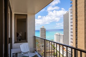 A balcony with a white chair, a small table, and a window with a view of the ocean and buildings.