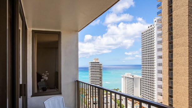 A balcony with a white chair, a small table, and a window with a view of the ocean and buildings.