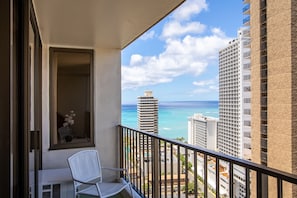 A balcony with a white chair, a small table, and a window with a view of the ocean and buildings. - Ocean View Condo | Aston Waikiki Banyan 1BPO3 – Pool & Hot Tub (Honolulu)