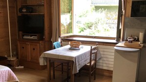L’intérieur d’un chalet accueillant, avec une table à manger en bois, des chaises, un téléviseur posé sur une étagère et une fenêtre donnant sur un paysage verdoyant.