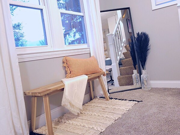 A room with a bench, a white rug, a staircase, and a mirror reflecting the staircase.