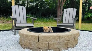 Twee Adirondack stoelen gericht op een ronde vuurkuip met daarin een boomstam, omringd door wit grind.