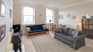 En stue med en blå sofa, en grå sofa, et spisebord med stole, en lysekrone og en bogreol.