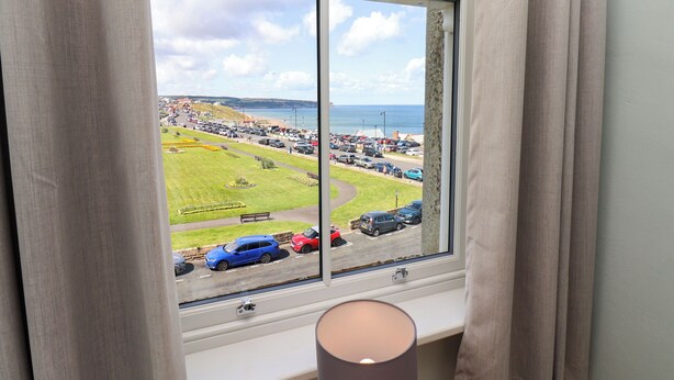 Et værelse med et stort vindue der kigger ud over en parkeringsplads og en græsplæne med en strand i det fjerne.