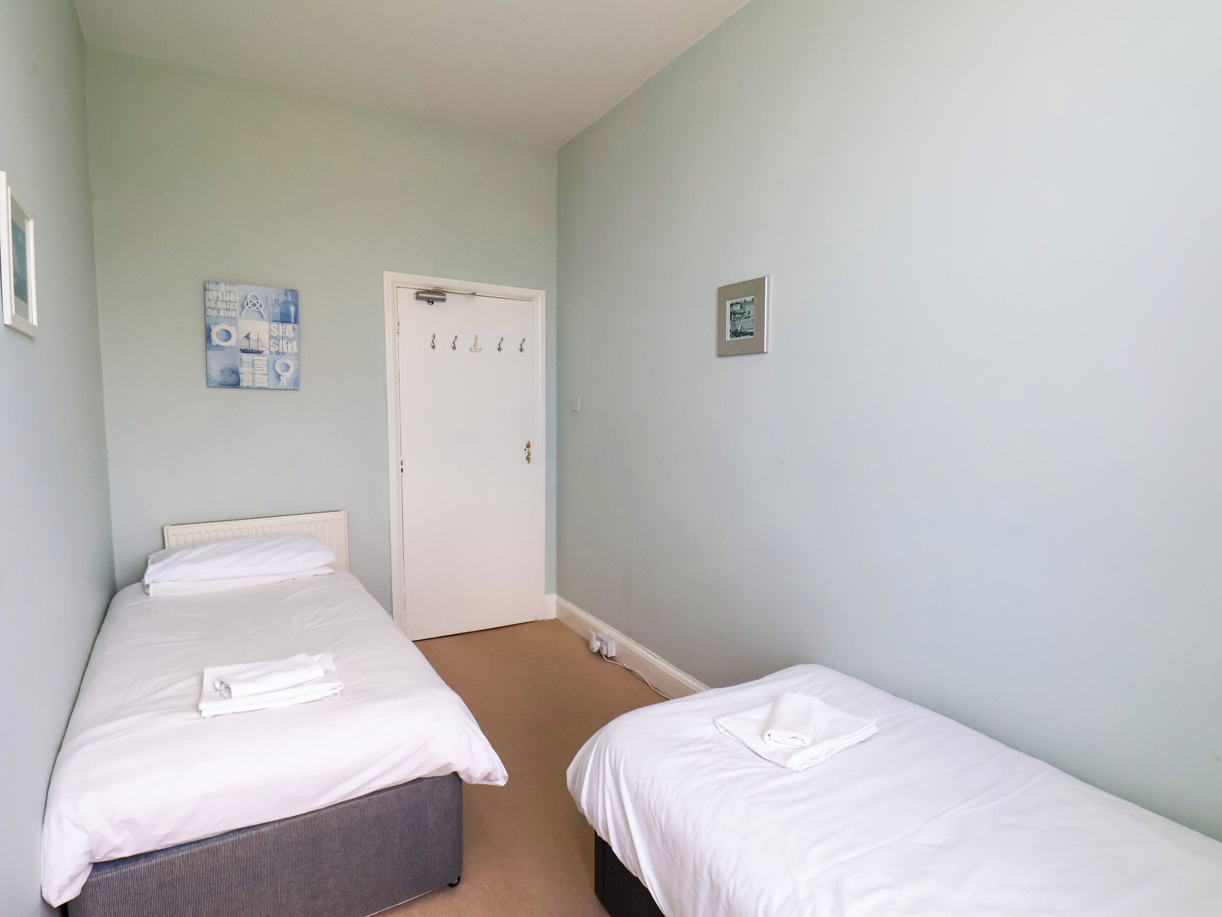 Una habitación con dos camas, una puerta con ganchos y un cuadro en la pared.