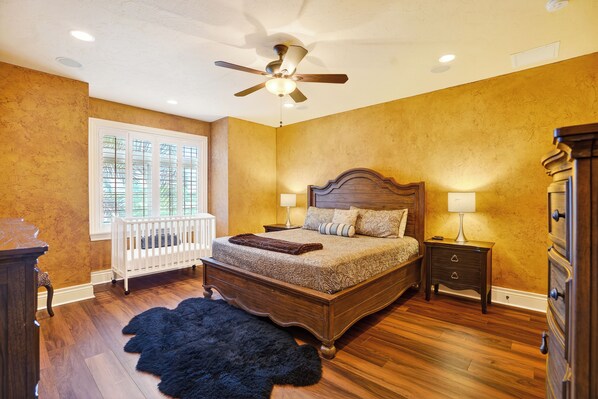 Une chambre à coucher avec un lit en bois, un ventilateur de plafond, une fenêtre avec des stores, une table de chevet et une commode.