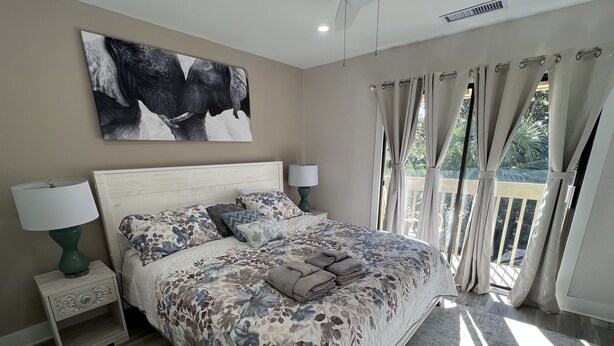 A bedroom with a large bed, floral bedding, a headboard, bedside tables with lamps, a ceiling fan, and a view of greenery through the window. - Spacious, cozy villa in Coligny Villas (Hilton Head Island)