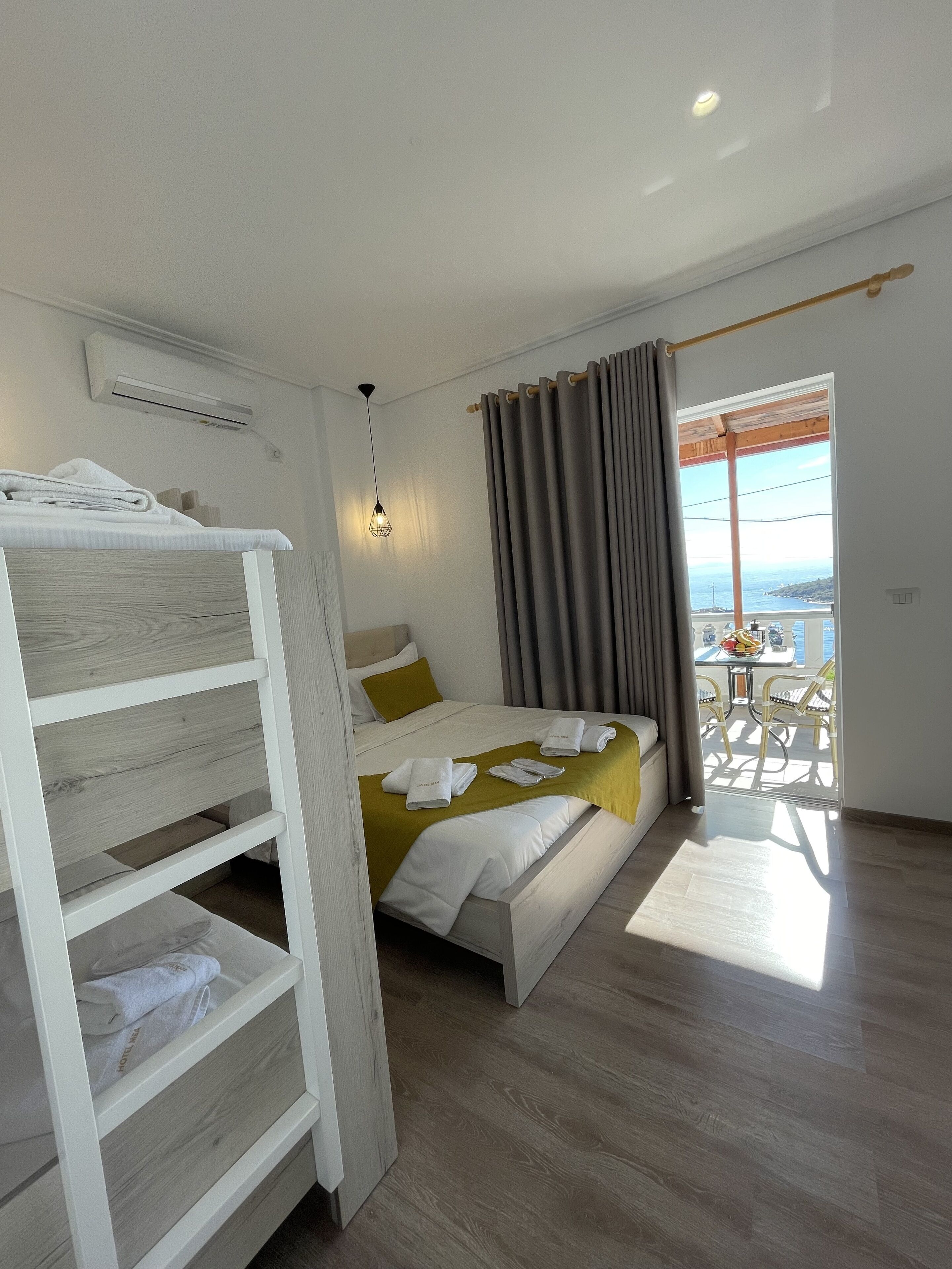 A hotel room with a bed, a small table with chairs, a balcony with a view, and a wooden bunk bed.
