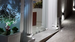 A modern building entrance with a glass door, white columns, and a small garden with a potted plant. - Relax Mea Hotel (Sarandë)