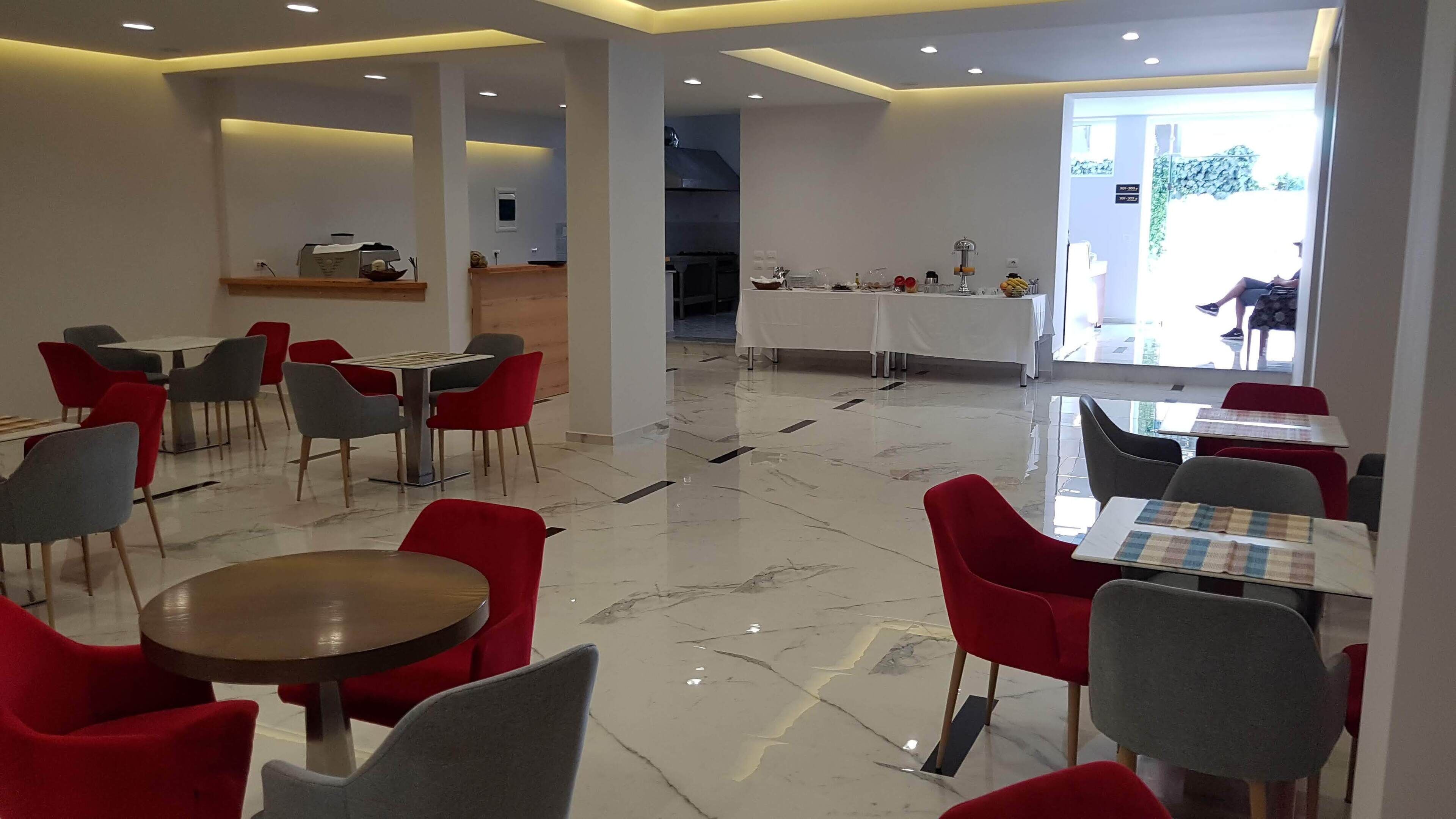 Un comedor moderno con sillas rojas y grises, mesas blancas y una cocina al fondo.