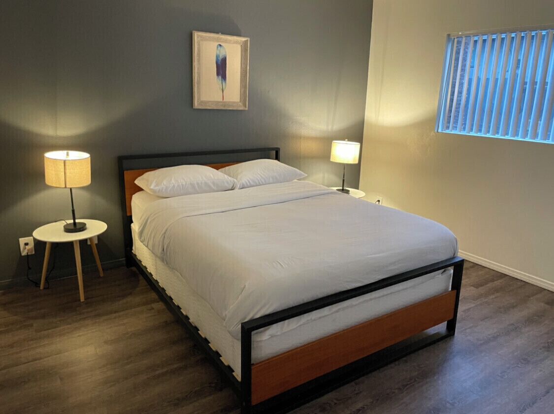 A neatly made bed with white linens in a room with a wooden floor, two bedside lamps, and a window with blinds.