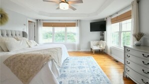 Une chambre à coucher comprenant un lit, une chaise, une commode, un téléviseur et de grandes fenêtres.