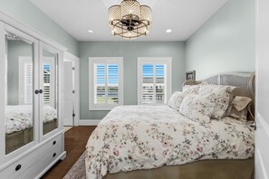 Une chambre avec un grand lit, une armoire, deux fenêtres avec volets et un lustre.