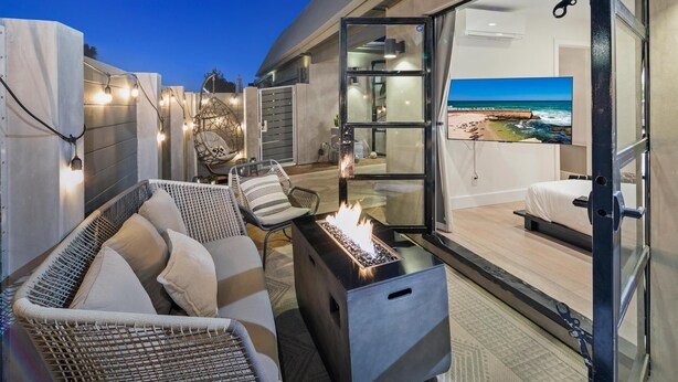 A modern outdoor seating area with a fire feature, wicker furniture, and a glass door leading to a bedroom with a beach view. - Ocean View Designer Retreat w/ EV Charger (San Diego)