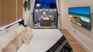 A bedroom with a bed, a TV, and a view of a balcony with outdoor seating and a fire feature. - Ocean View Designer Retreat w/ EV Charger (San Diego)