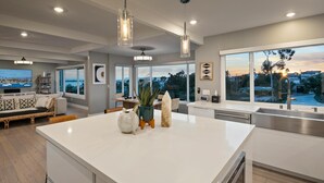 A modern kitchen with a large island, a dining area with a sofa, and a view of the cityscape through large windows. - Ocean View Designer Retreat w/ EV Charger (San Diego)
