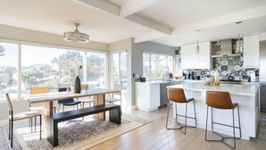 A modern kitchen with a large island, bar stools, and a dining area with a bench and chairs. - Ocean View Designer Retreat w/ EV Charger (San Diego)