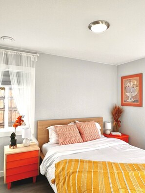 Une chambre à coucher avec un lit, une table de chevet, une lampe, un vase avec des fleurs et une fenêtre ornée de rideaux légers.