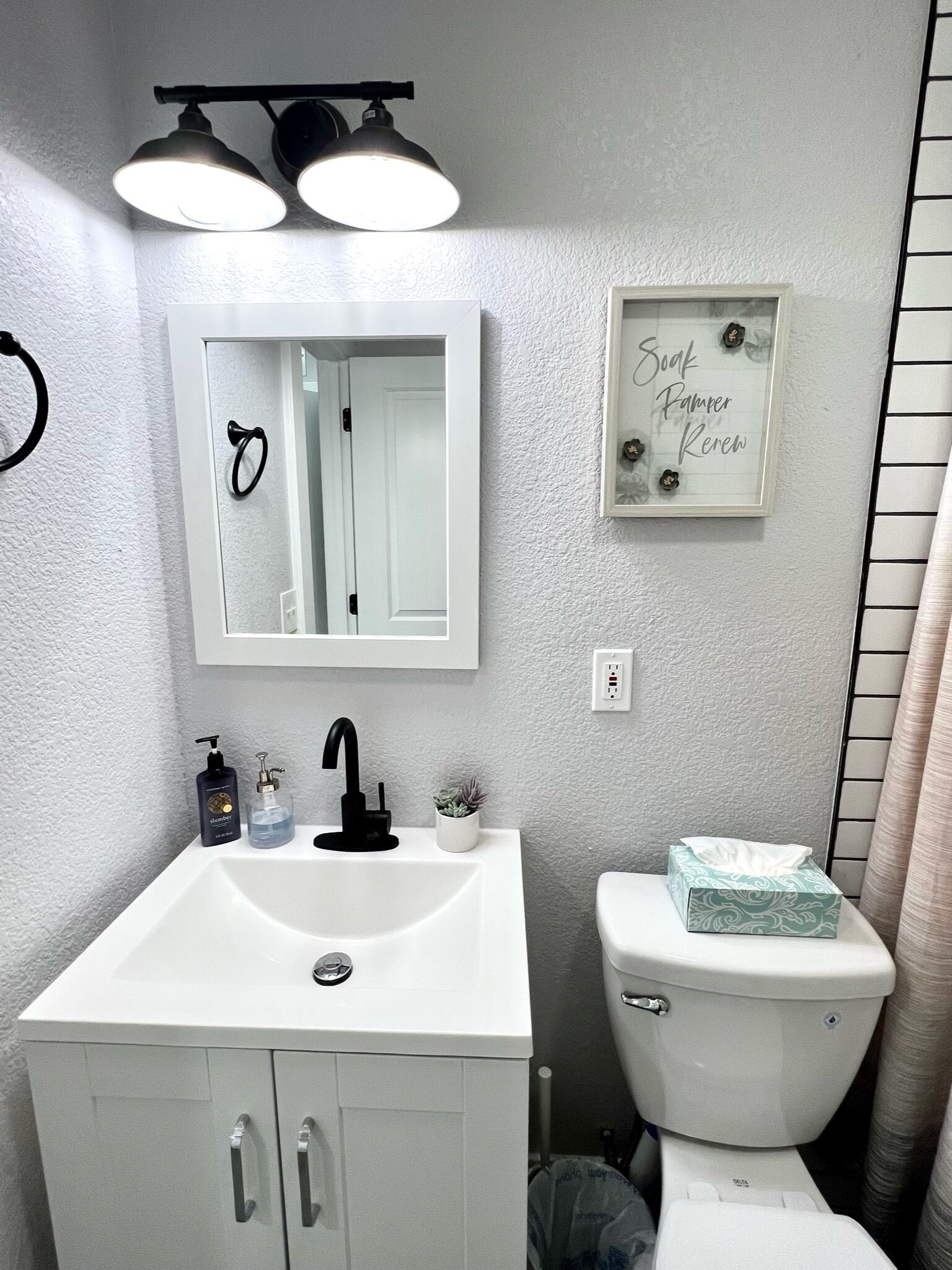 A bathroom with a white sink, black faucet, and a mirror above it. There is a framed sign on the wall and a toilet with a tissue box beside it.