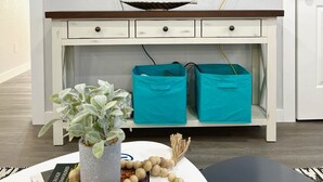A living room with a flat-screen TV mounted on a white wall, a wooden console table with drawers, two teal storage bins, a potted plant, and a decorative garland.