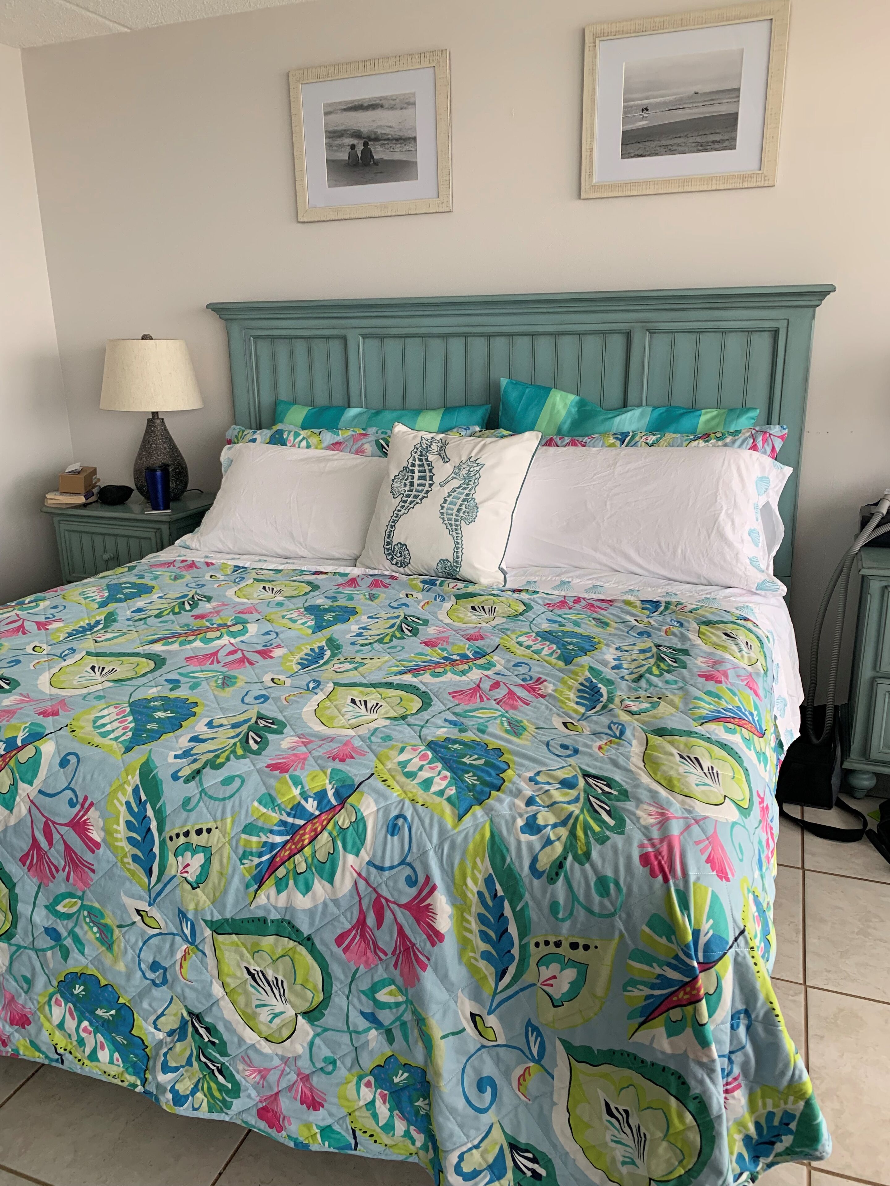 Uma cama com colcha floral colorida, cabeceira verde-água e duas fotografias emolduradas na parede.