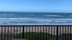 Vista a la playa desde un balcón con barandilla, olas del océano y una playa de arena.