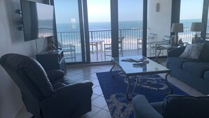 Sala de estar con televisor grande, sofá, sillón y una mesa. Hay un balcón con vista al océano.
