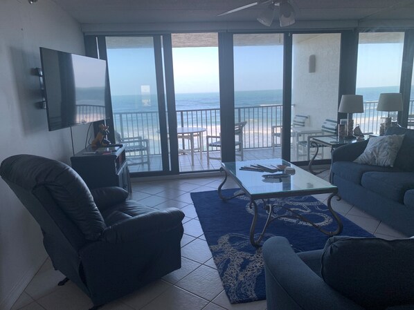 A living room with a large TV, a sofa, a chair, and a table. There is a balcony with a view of the ocean.