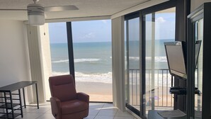Habitación de hotel con un gran ventanal corredizo a la playa, una silla roja, una cama con una manta colorida y un escritorio con televisor de pantalla plana.
