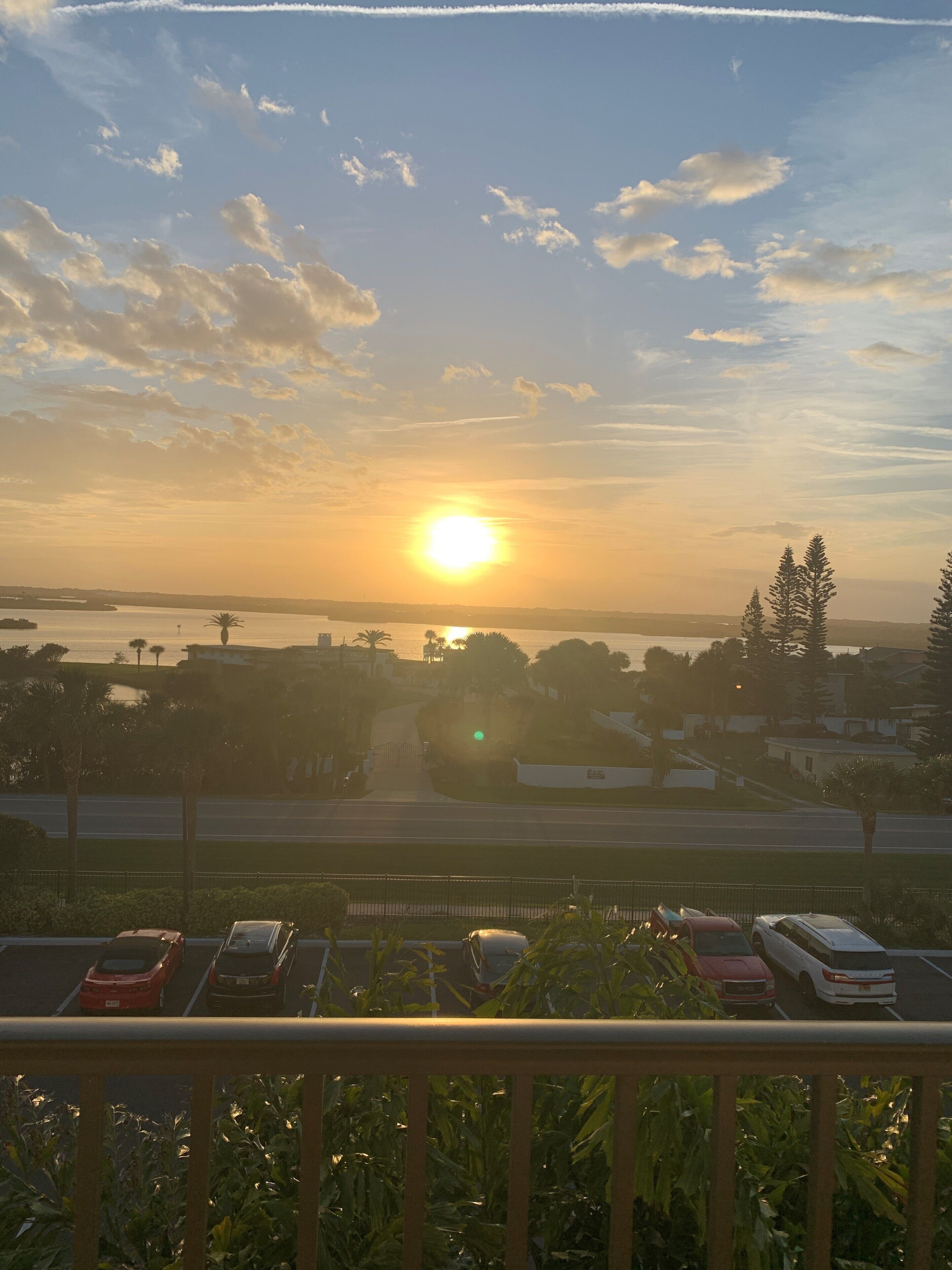 Vista del atardecer desde un balcón con vista a un estacionamiento, coches y un cuerpo de agua al fondo.