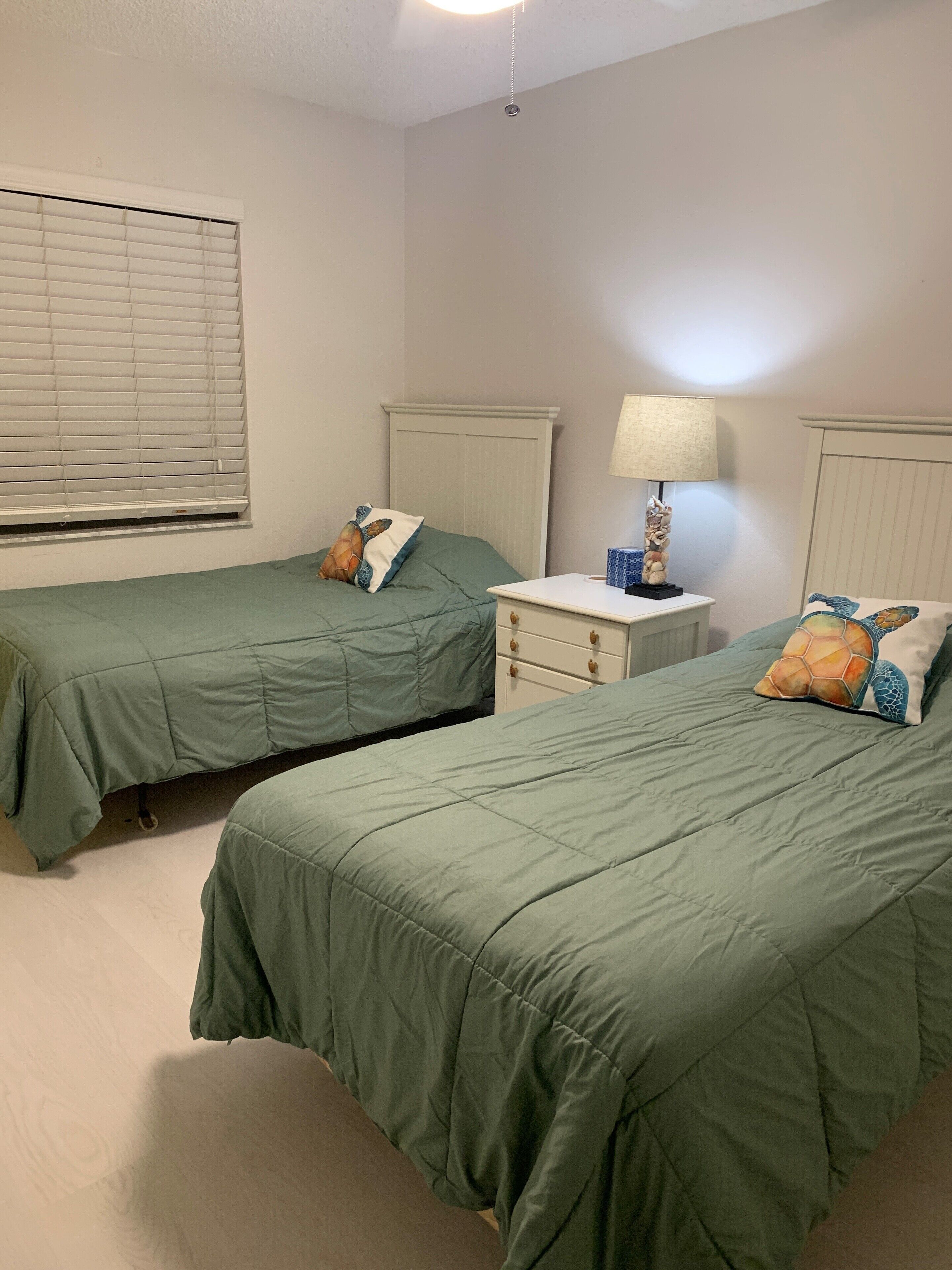 Un dormitorio con dos camas individuales, una mesita de noche con lámpara y una ventana con persianas.