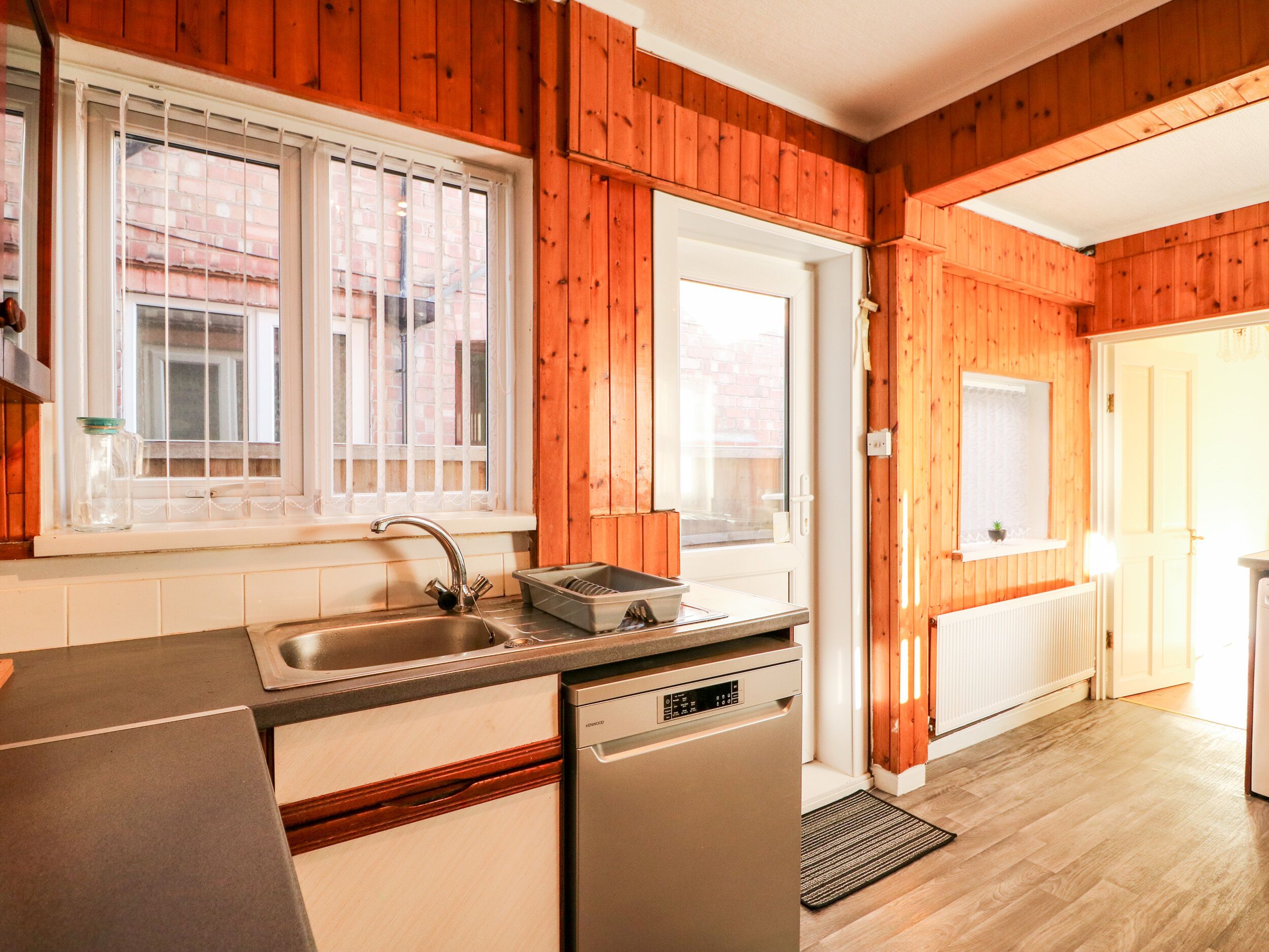 A kitchen with wooden paneling, a sink, a dishwasher, and a window with blinds.