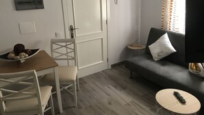 Un petit salon moderne avec un canapé gris, une table à manger en bois avec des chaises, une petite table ronde avec une télécommande, et une porte avec une poignée.
