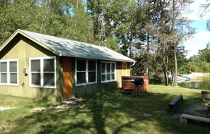 Situated in a wooded area, this cabin features a front porch, a wooden deck, and a small grill, offering scenic views of a nearby body of water.