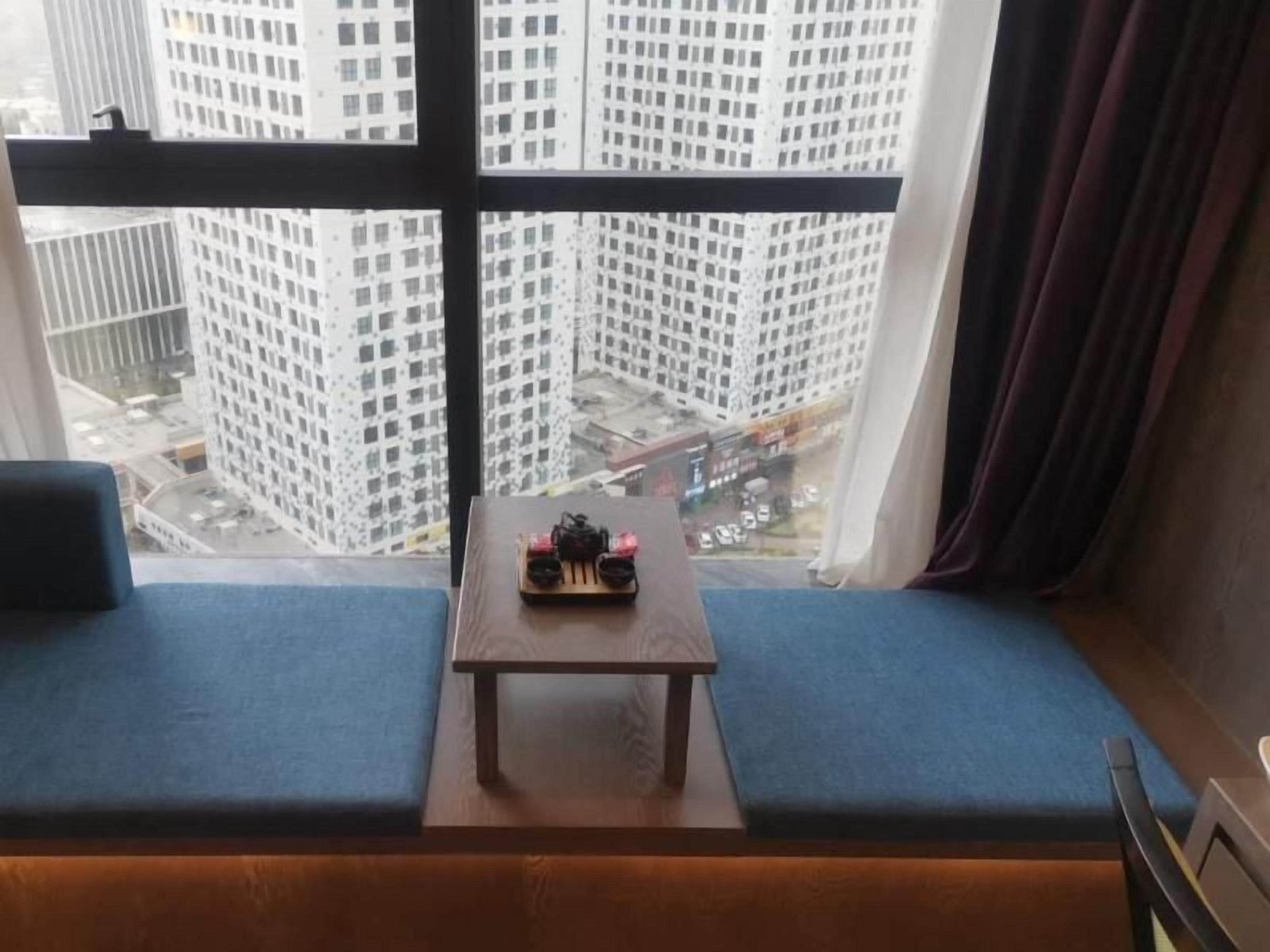 A room with a view of tall buildings through a window, a small table with a decorative item, and a blue cushioned bench.