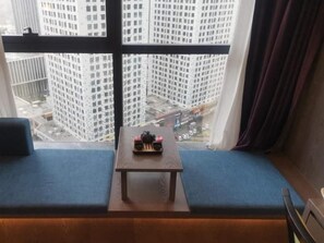 Ein Zimmer mit Blick auf hohe Gebäude durch ein Fenster, ein kleiner Tisch mit einem dekorativen Gegenstand und eine blaue gepolsterte Bank.
